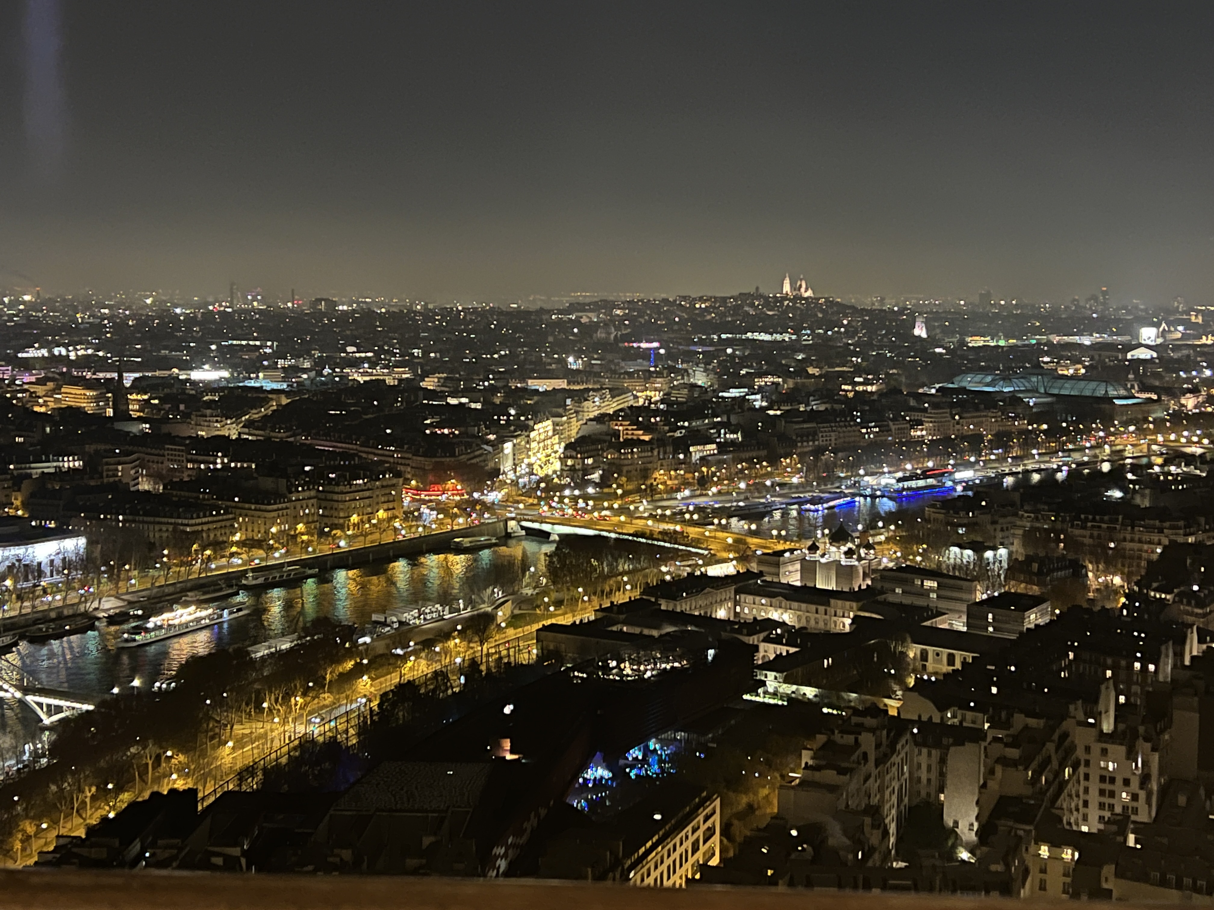A view of Paris lit up at night. The river Seine is prominent in the foreground, with lights reflecting on its surface.