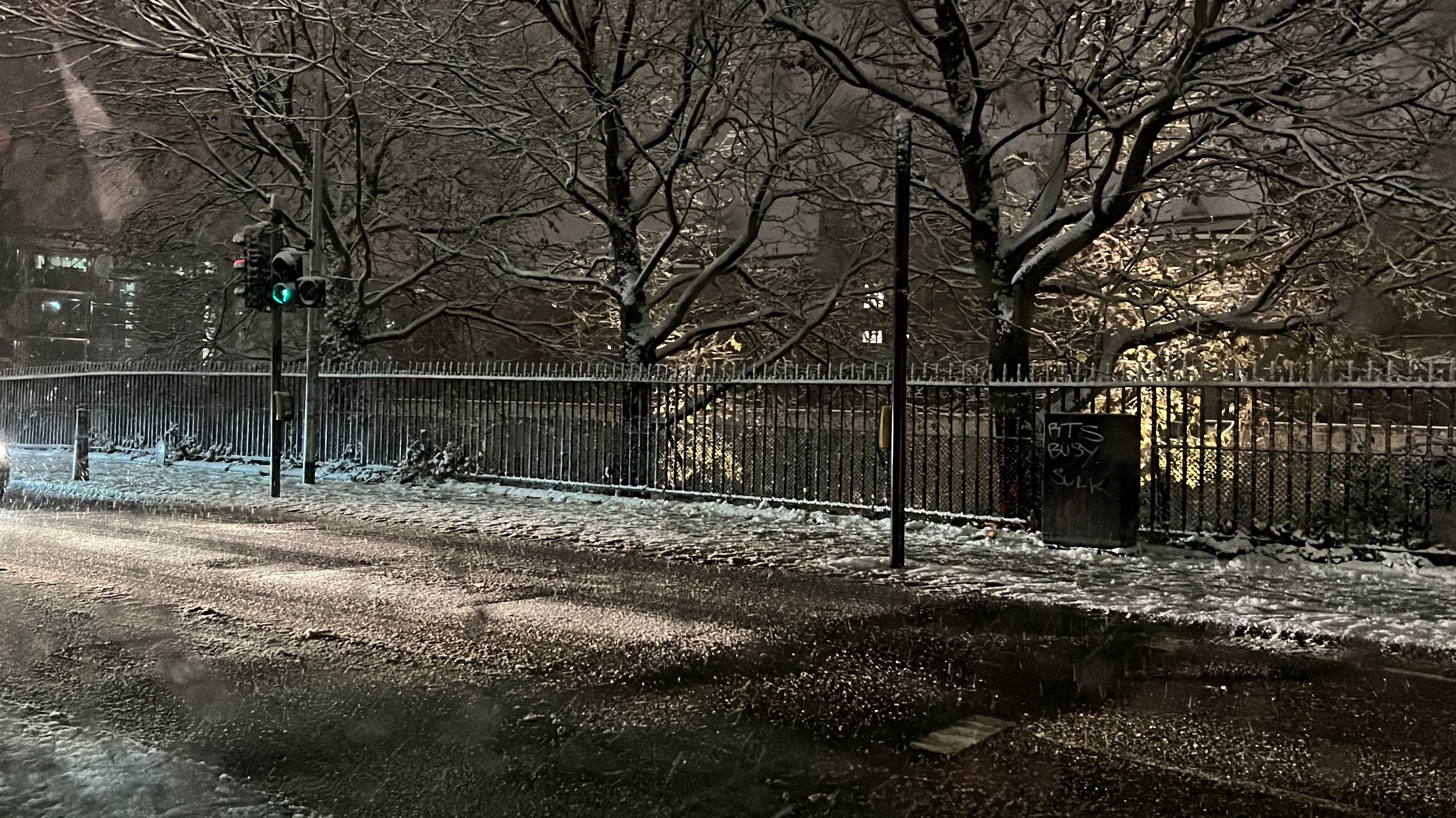 Image of a street and sidewalk lightly covered with snow. On the other side of the street is a wrought iron fence and some trees.
